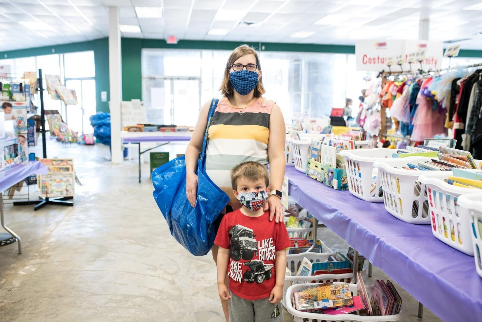 A mom and her four-year-old son stand with their masks and shopping bag in front of baskets of books and costumes.