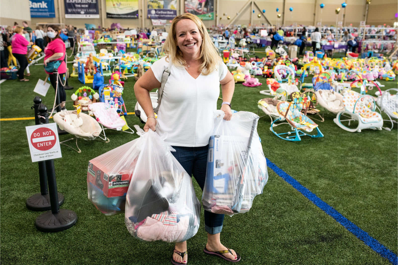 A mom with a large JBF shopping bag on her shoulder stands beside her husband who wears their toddler at a JBF sale.